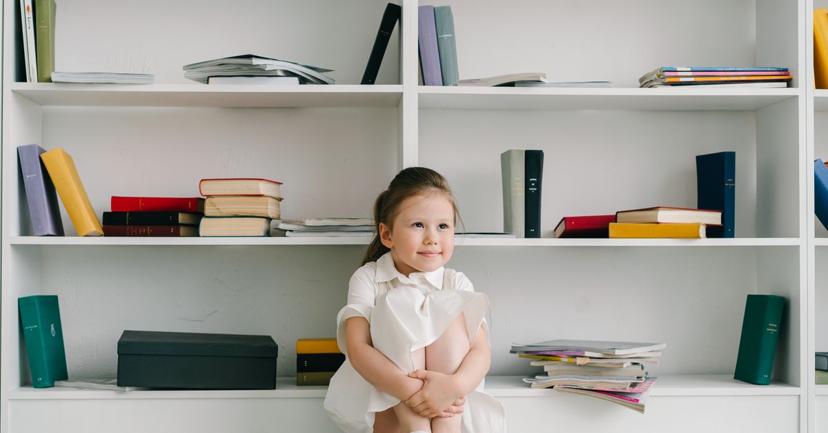 A cute child sitting on the floor by a bookshelf, surrounded by books in a cozy indoor setting.