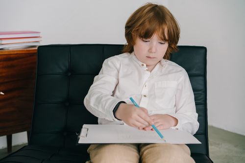 Free Child with red hair concentrates on drawing with colored pencils while sitting on a black chair. Stock Photo