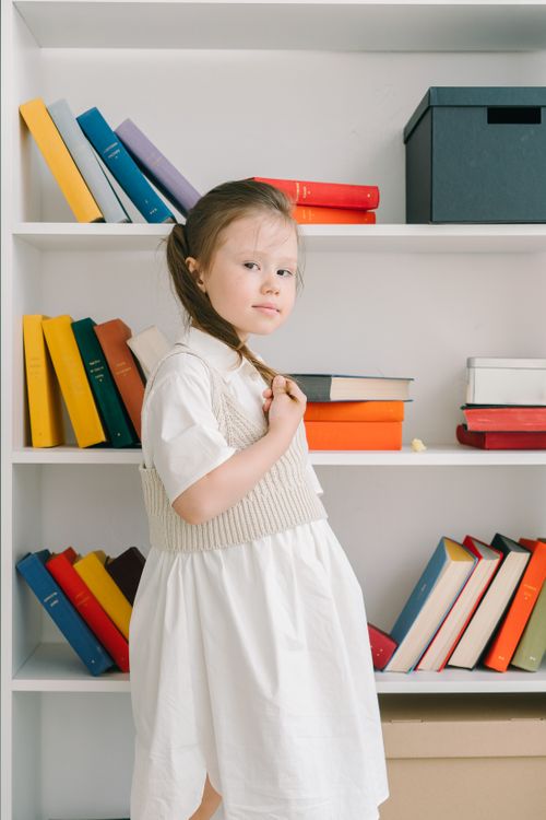 Free A young girl with braided hair in a white dress stands by a colorful bookshelf indoors. Stock Photo