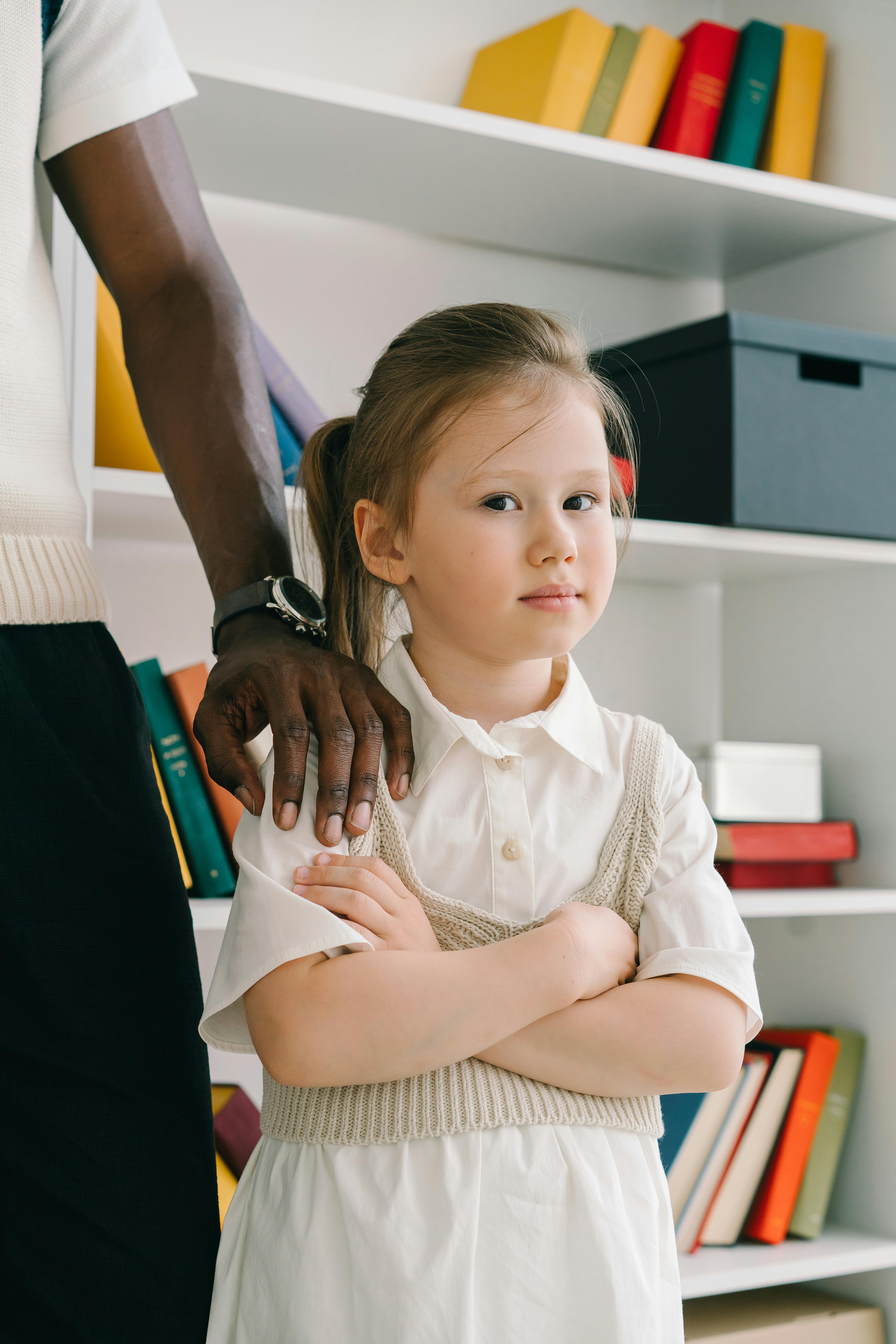 Young girl stands with arms crossed, thoughtful in a cozy library.