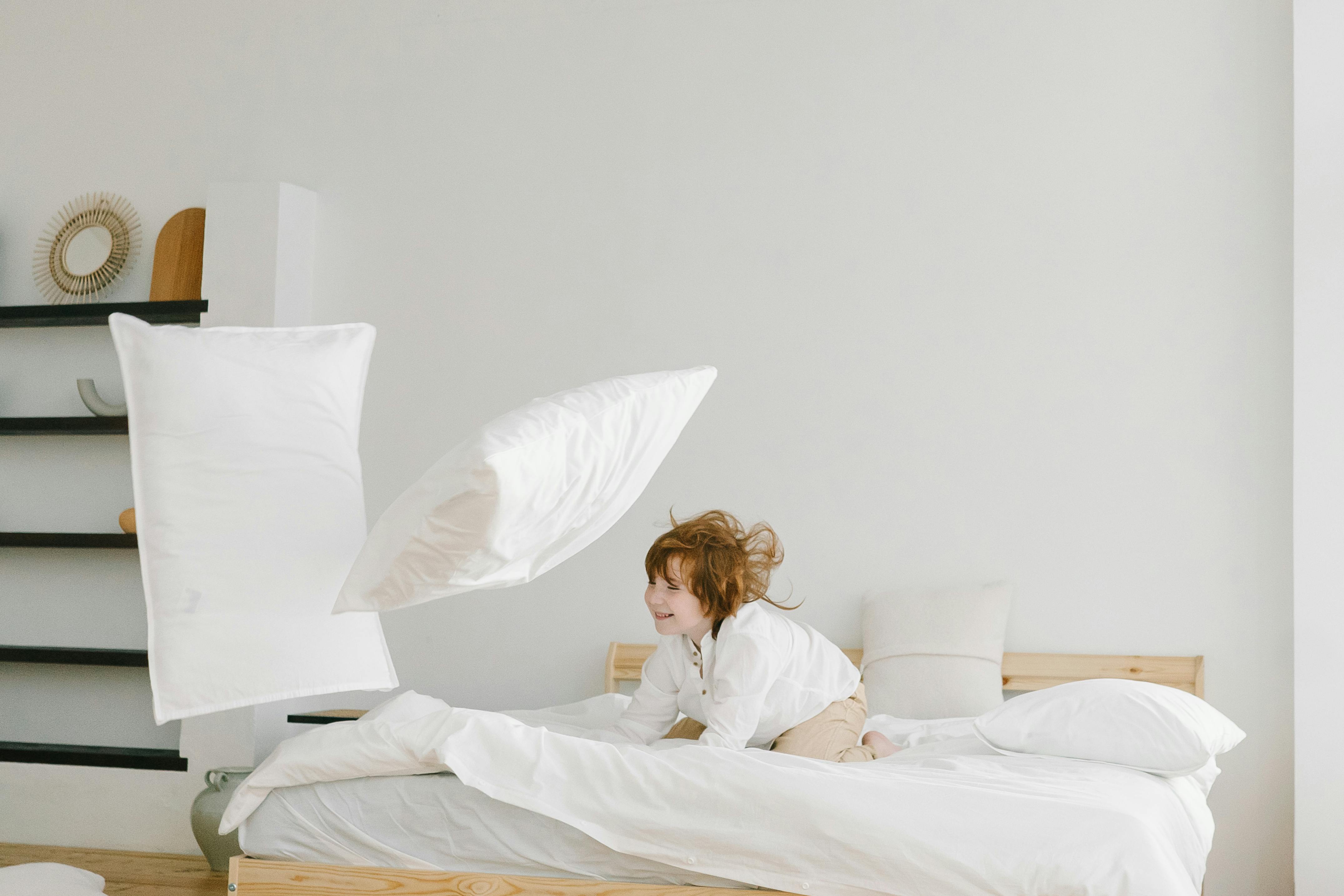 A joyful child playing and throwing pillows in a bright, minimalist bedroom setting.