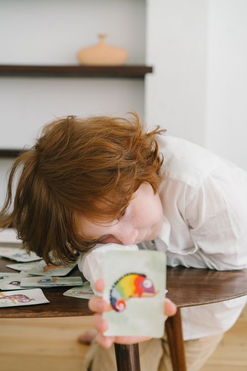 Free A redheaded child engaging in a playful pose with a colorful chameleon-themed card indoors. Stock Photo