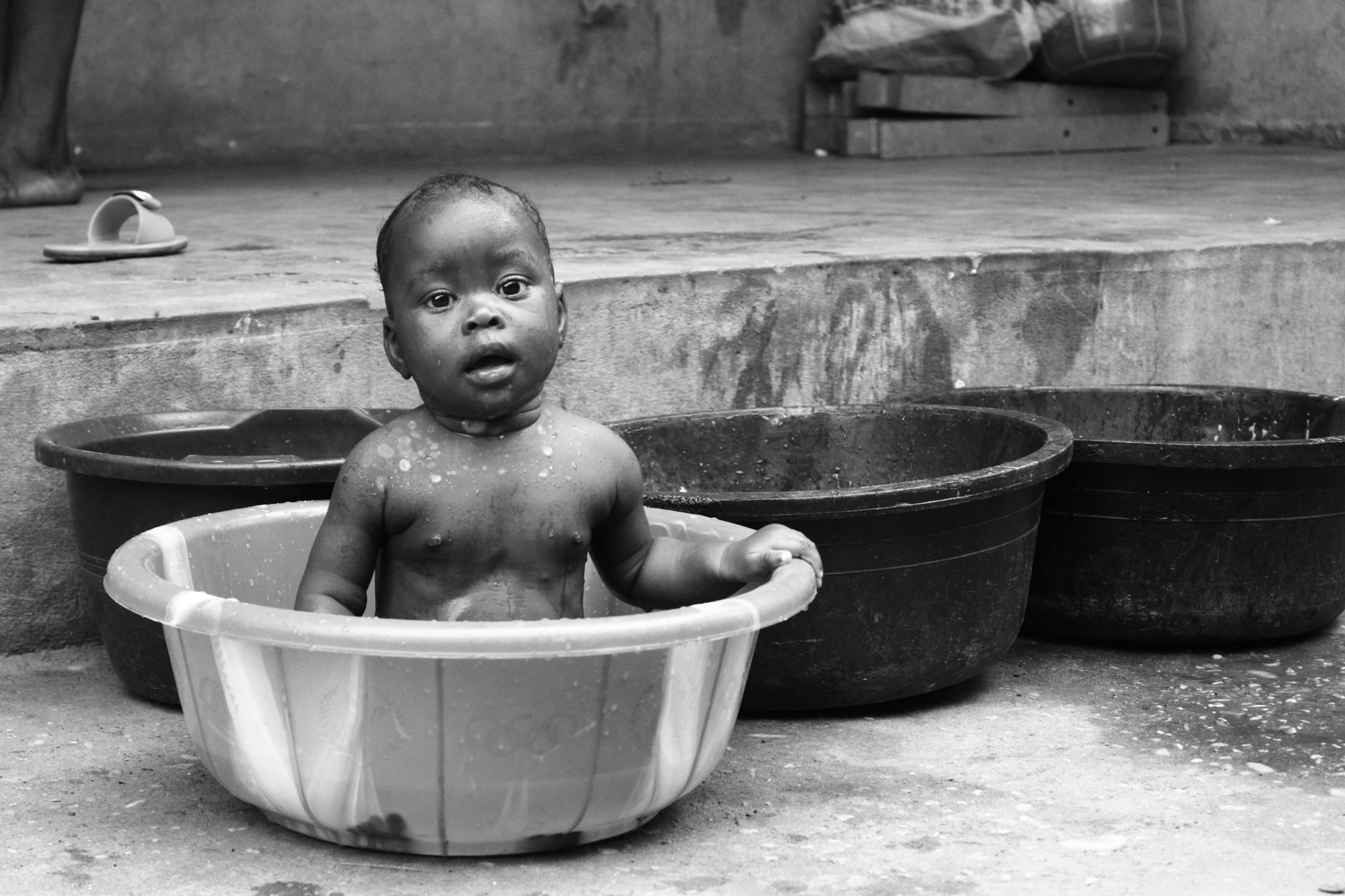Charming black and white photo of a baby bathing in a basin outdoors in Cotonou, Benin.