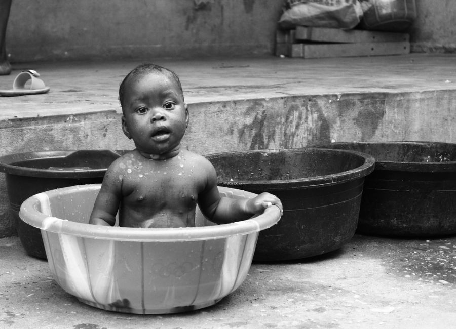 Charming black and white photo of a baby bathing in a basin outdoors in Cotonou, Benin.