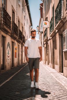 A bearded man in a white shirt strolls through a European street bathed in sunlight, depicting a relaxed atmosphere.