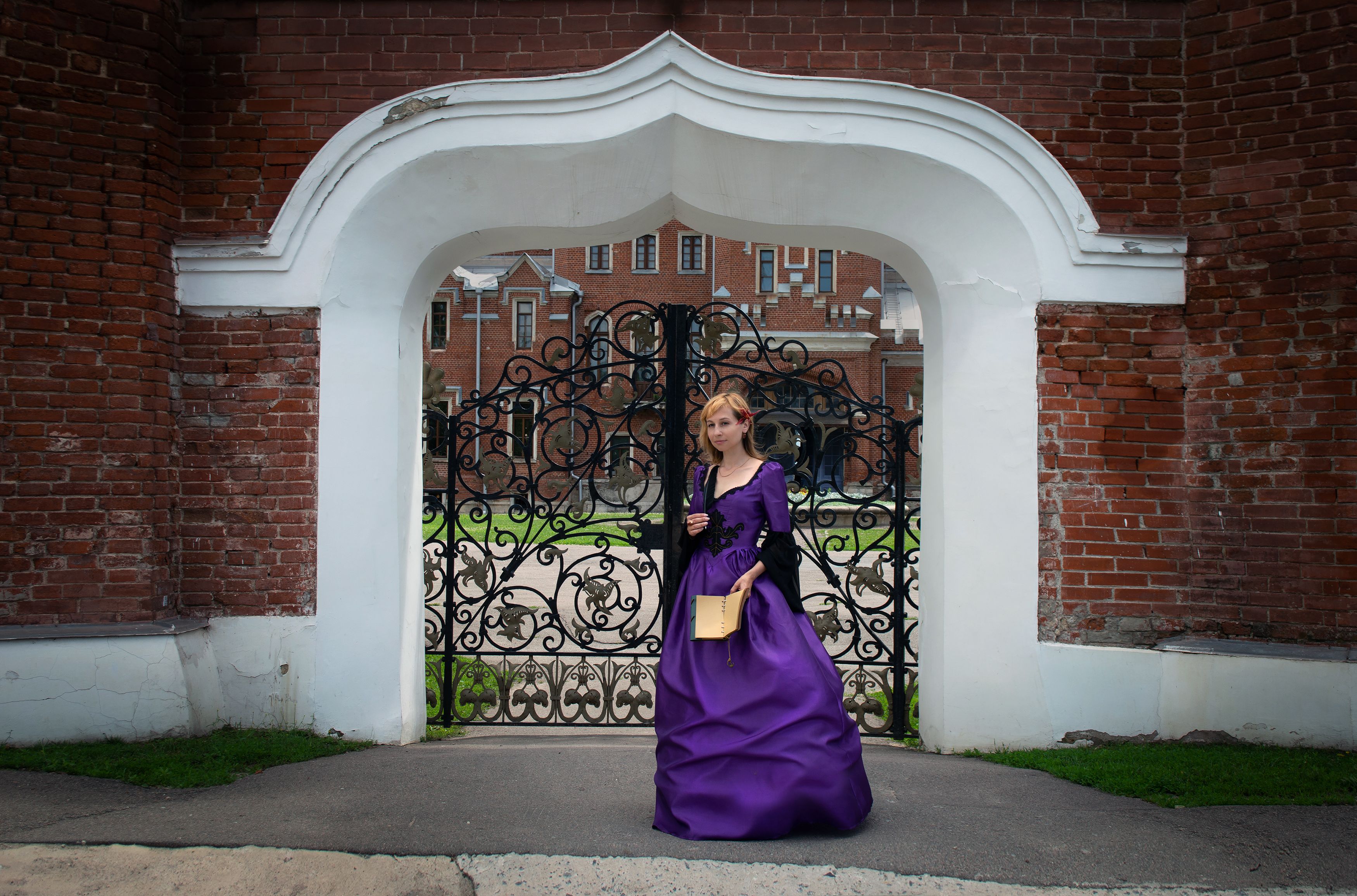 A woman in a purple gown stands elegantly by a historical wrought iron gate framed by brick and arch architecture.
