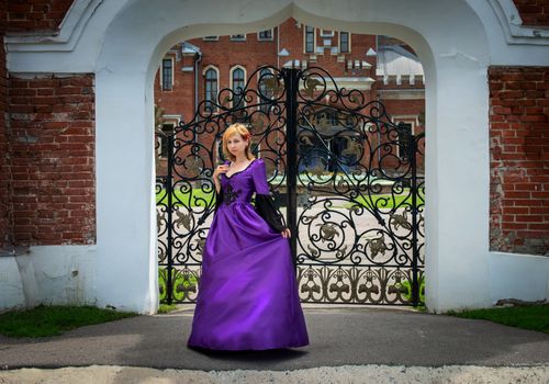 Woman in a vibrant purple dress poses elegantly by decorative wrought iron gates in a historic setting.