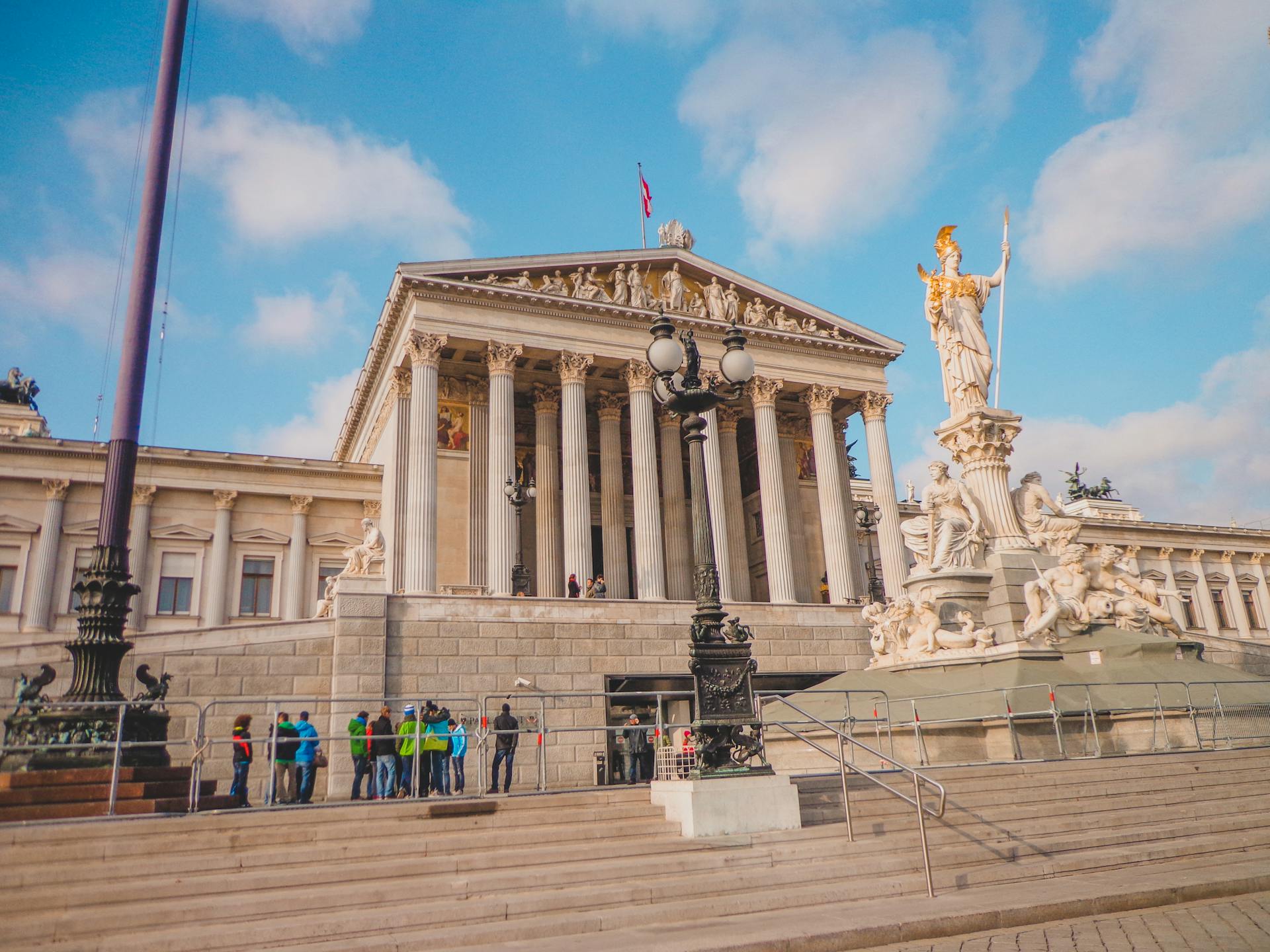 Exterior view of the Vienna Parliament Building with iconic Athena Fountain and tourists.