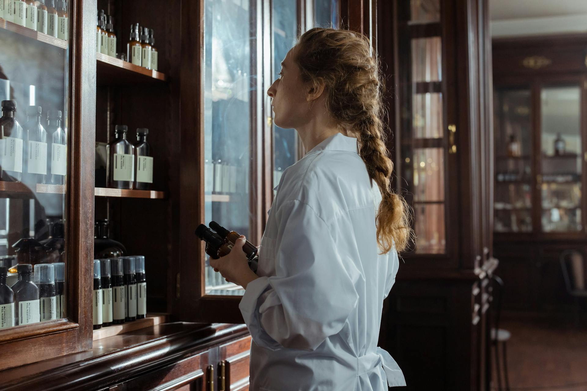 Woman in white coat examines medicine bottles in a vintage pharmacy setting.