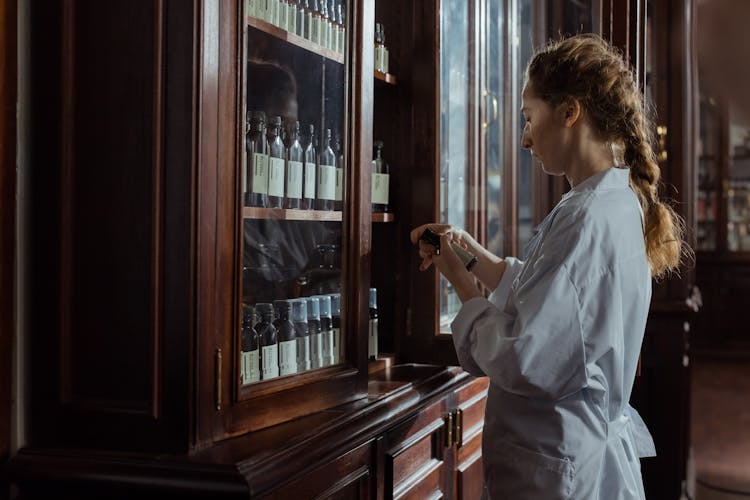 Woman In White Uniform Looking At The Product She Is Holding From The Cabinet