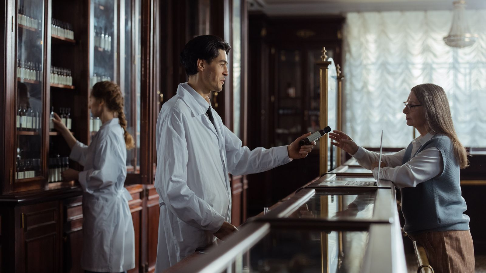 A pharmacist hands medication to a customer at a classic vintage pharmacy counter.