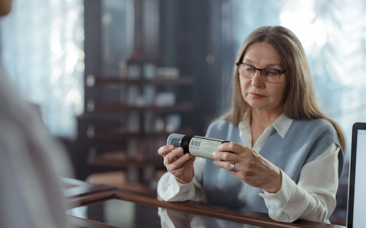 A senior woman with eyeglasses reviews a medicine bottle at a pharmacy counter indoors.
