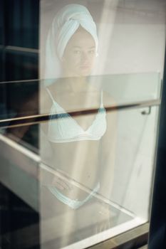Artistic reflection of a woman in bikini standing indoors by a glass window.