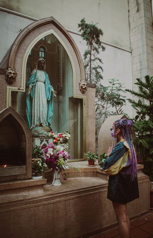 Free A woman with colorful hair prays in a chapel before a Virgin Mary statue surrounded by flowers. Stock Photo