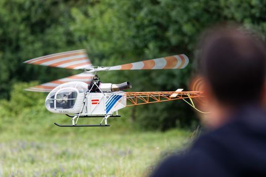 A remote-controlled helicopter flies outdoors with a person in the background, focusing on its rotors.