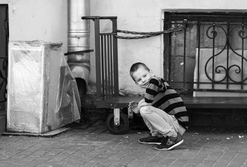 Grayscale Photo of Boy in Stripe Shirt Sitting on the Street