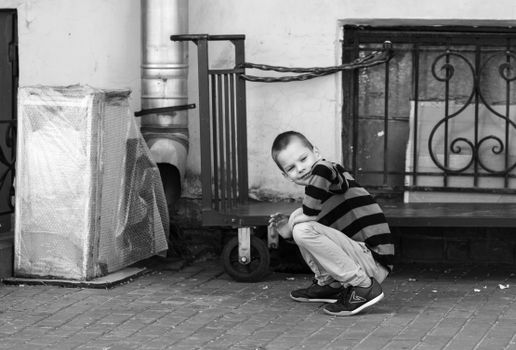 A young boy crouching beside a cart on the pavement, captured in monochrome.