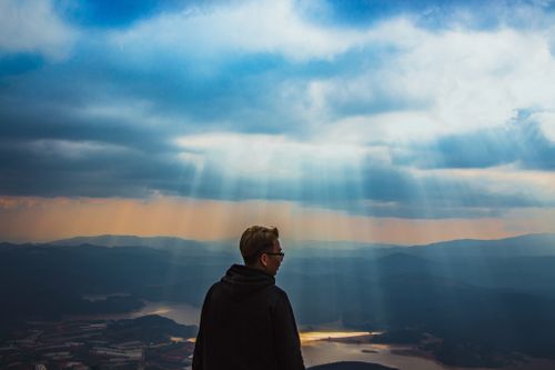 Free A man stands on a hill, admiring sunlight breaking through clouds over scenic landscape. Stock Photo
