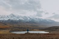 Person in Blue Jacket Standing on Brown Grass Field Near Lake and Snow Covered Mountains during