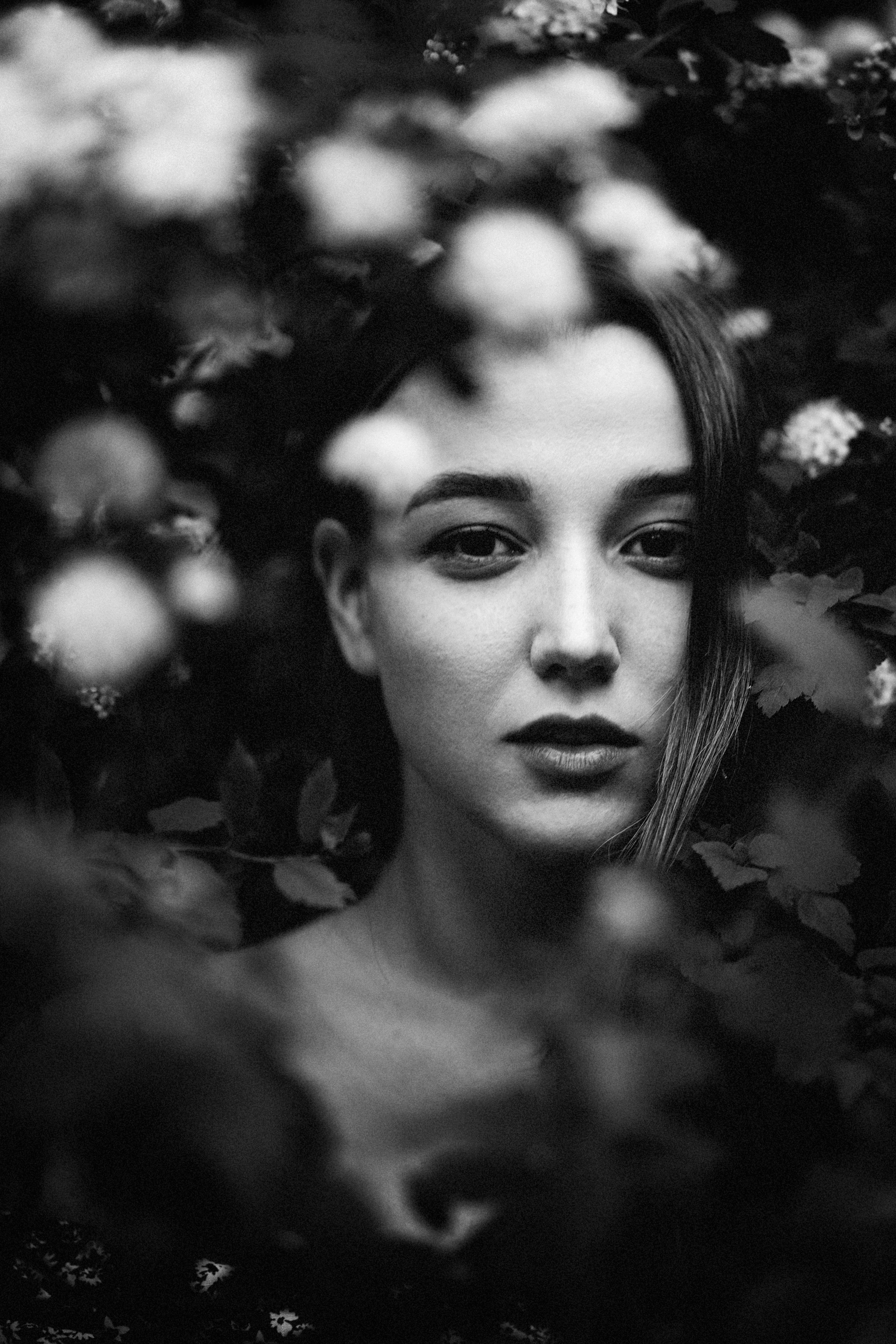 Black and white portrait of a woman surrounded by blurred foliage, evoking a mysterious aura.