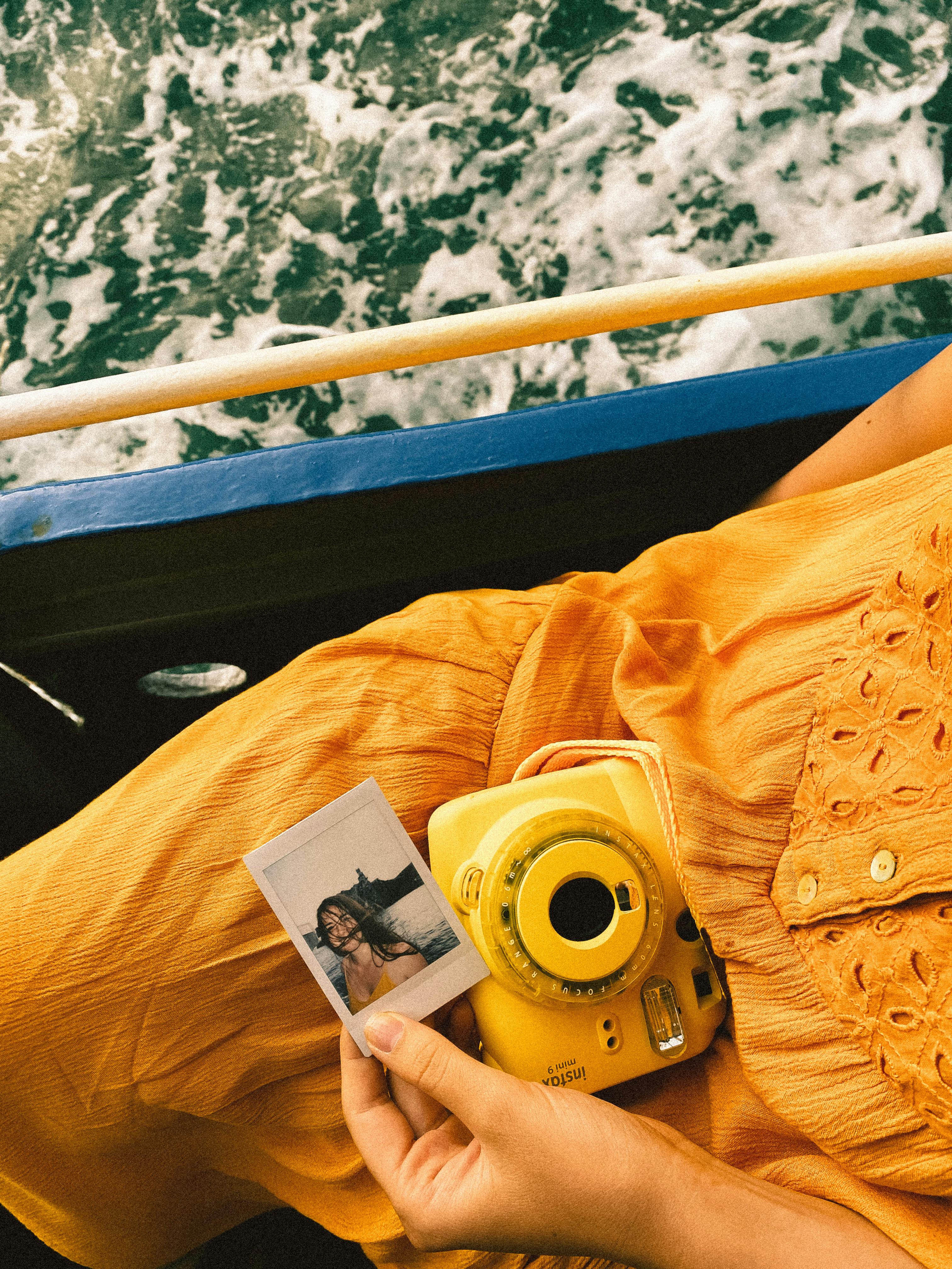 A hand holding a Polaroid photo and yellow instant camera on a boat ride over water.