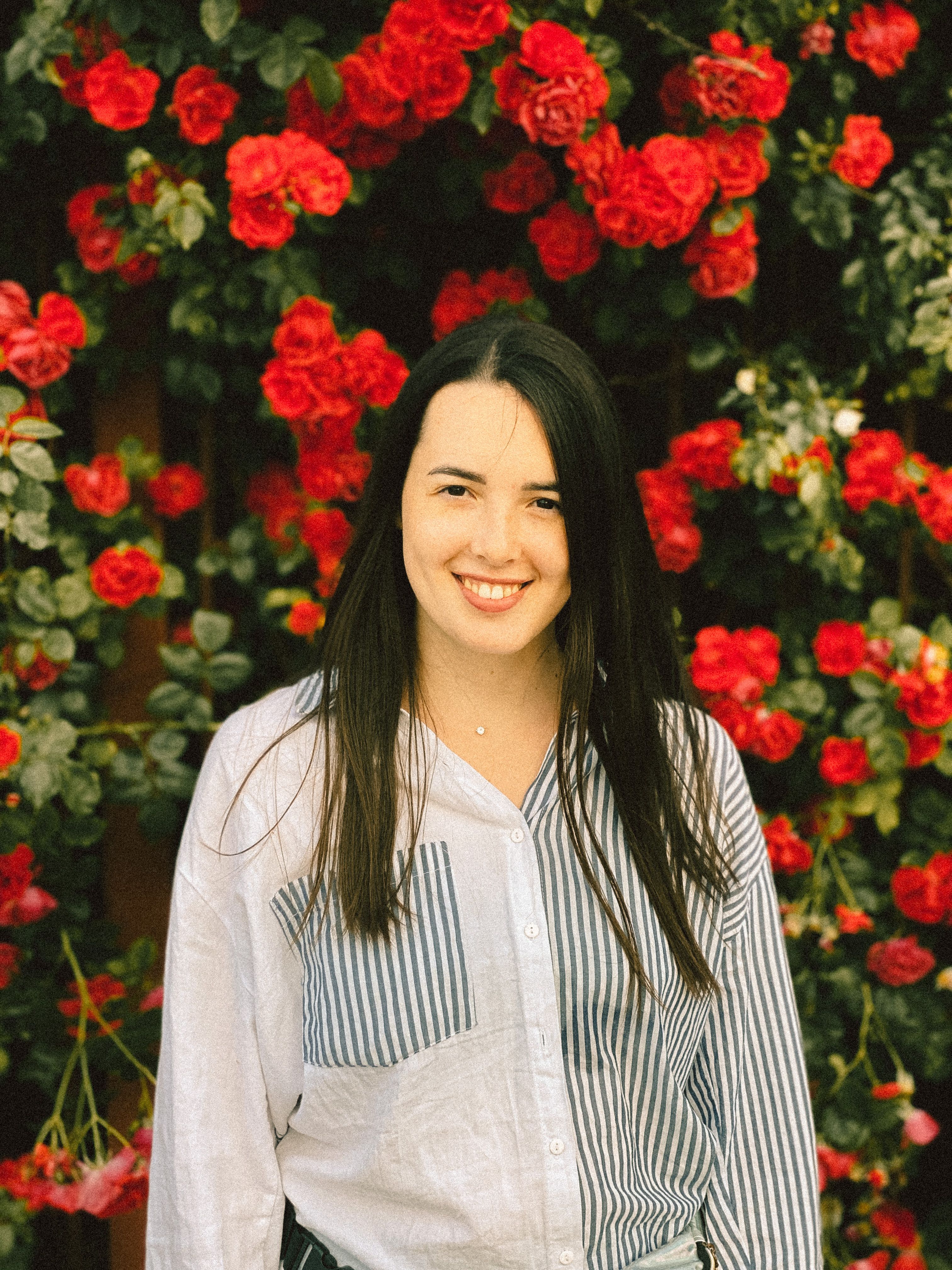 Bright portrait of a smiling woman standing in front of vibrant red roses in a garden setting.