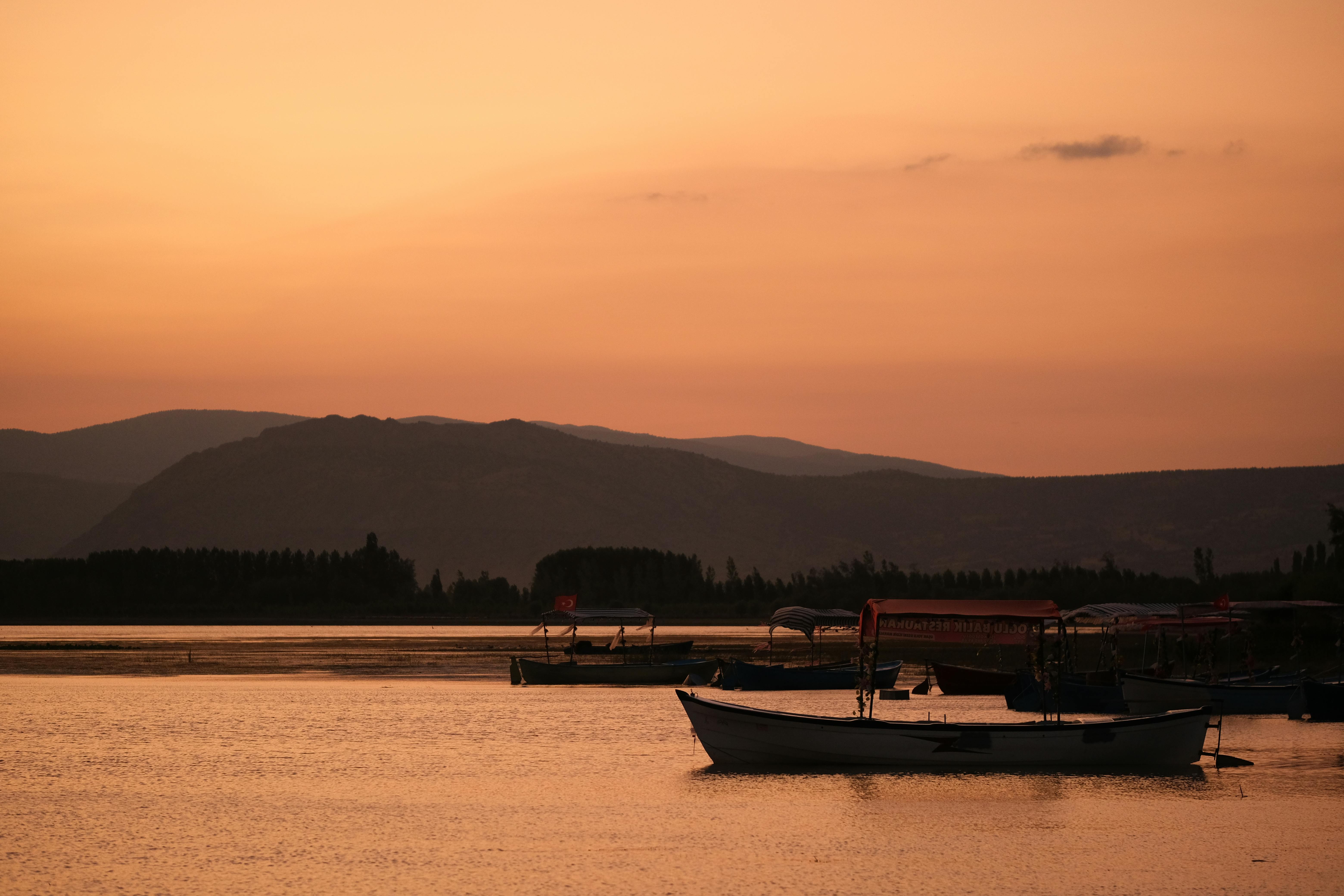 Free Tranquil scene of boats on calm water under a vibrant sunset with mountain backdrop. Stock Photo