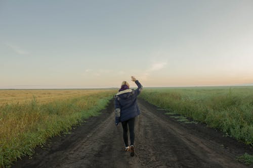 Person Wearing Blue Puffer Jacket Walking on the Road Between Green Grass