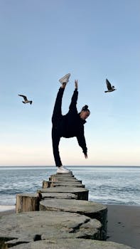 A person performs a dance pose on beach posts, surrounded by flying seagulls by the ocean.
