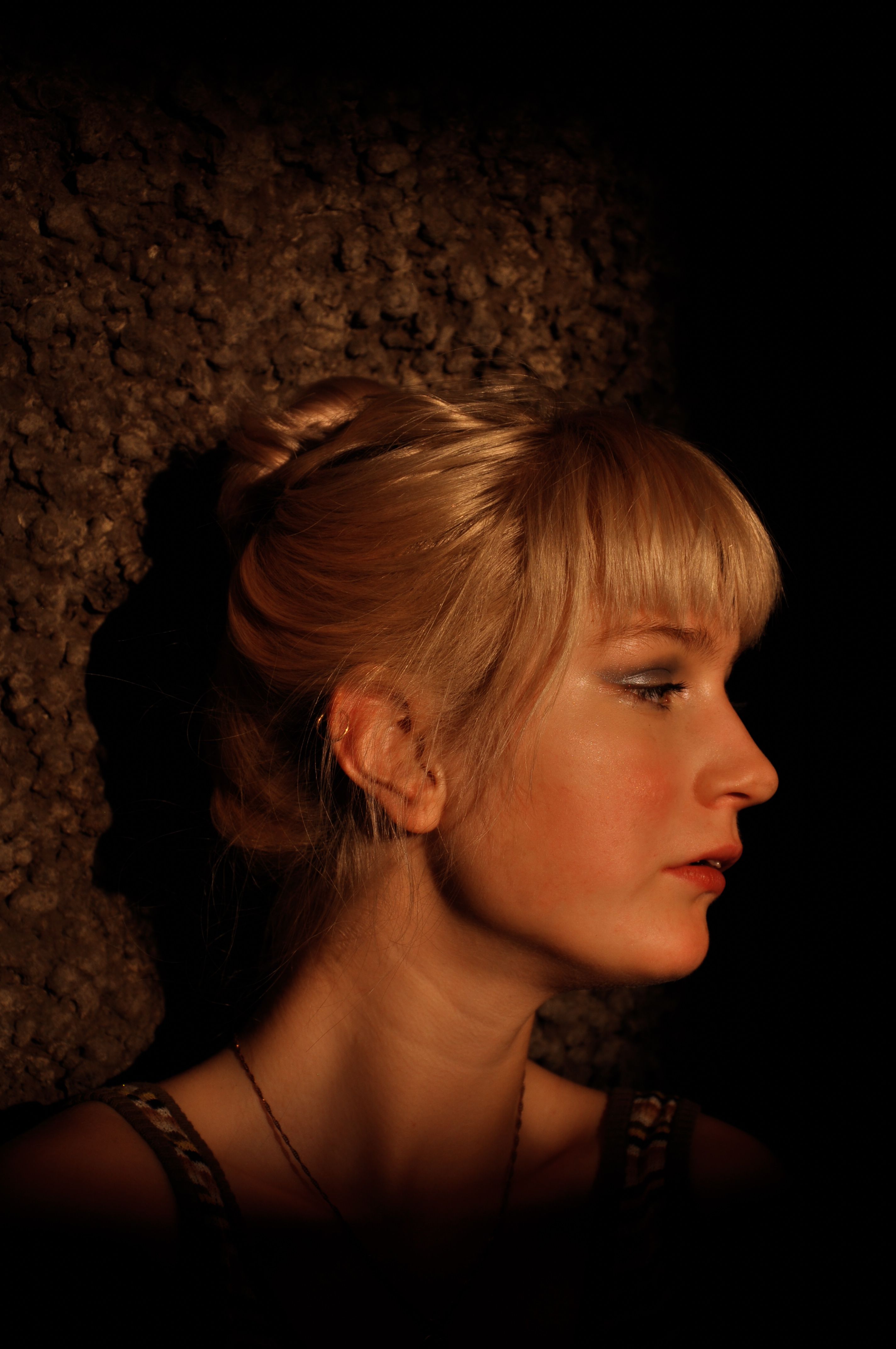 Portrait of a young woman with blonde hair against a dark textured background, side view, dim lighting.