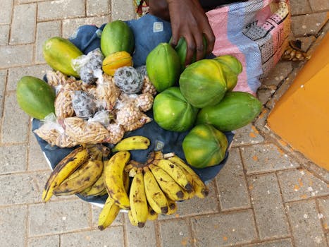 A variety of tropical fruits and nuts neatly arranged on a market stall, showcasing papayas, bananas, and bags of mixed nuts.