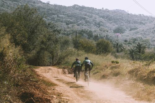 Free Cyclists biking through a dusty trail surrounded by lush hills in Brazil on a sunny day. Stock Photo