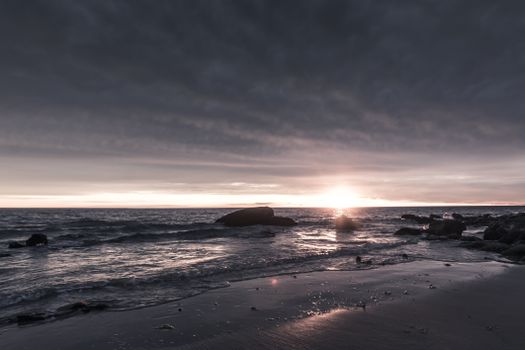 A dramatic seascape with dark clouds and a stunning sunset over a rocky shore.
