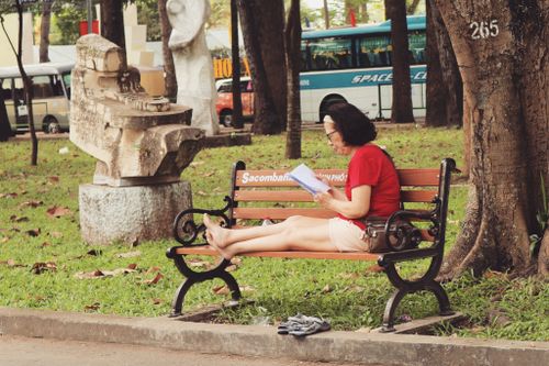 Free Woman relaxed on bench reading a book in an outdoor park setting with urban background. Stock Photo