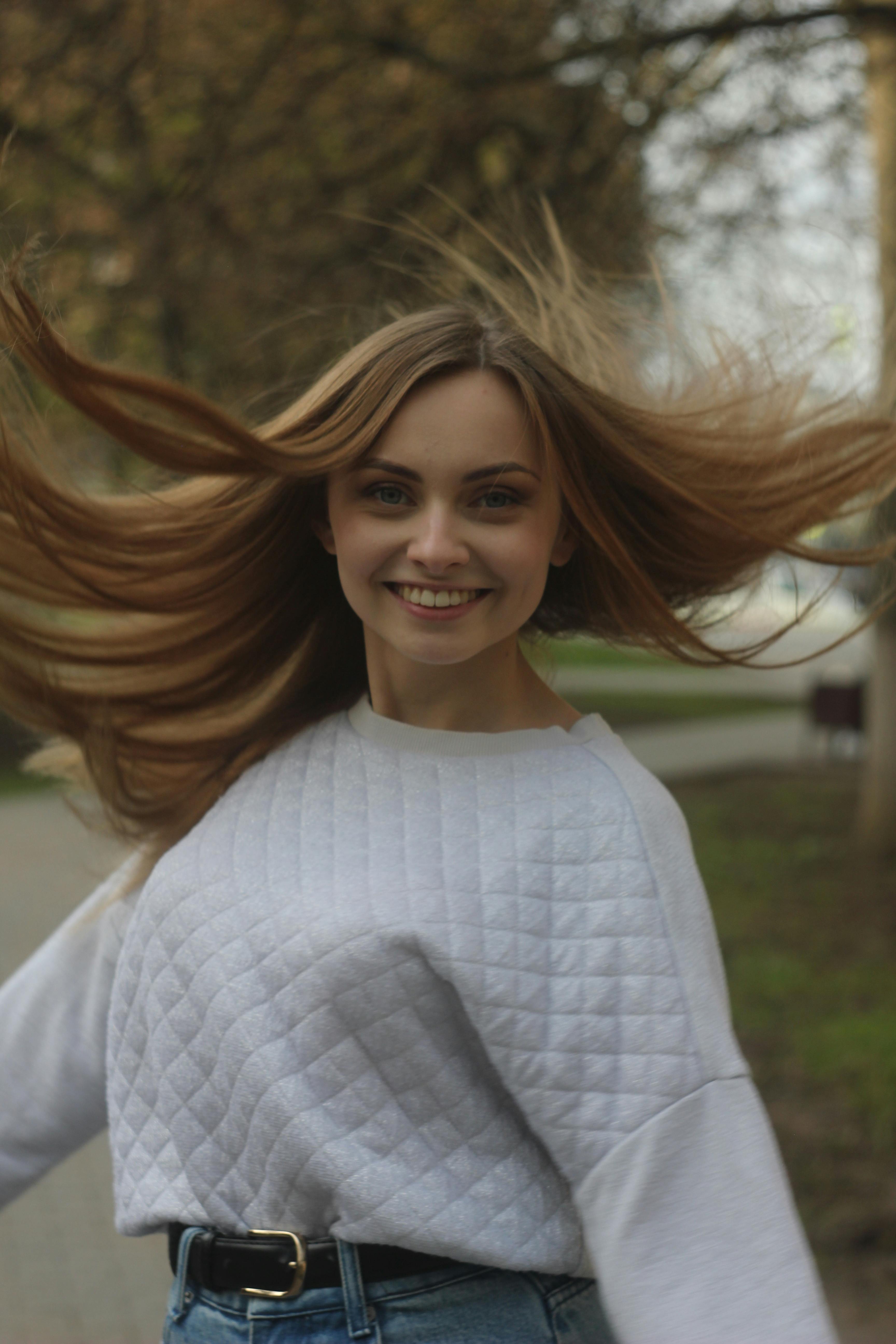 Smiling woman in white sweater spinning joyfully outdoors with flowing hair and trees in the background.