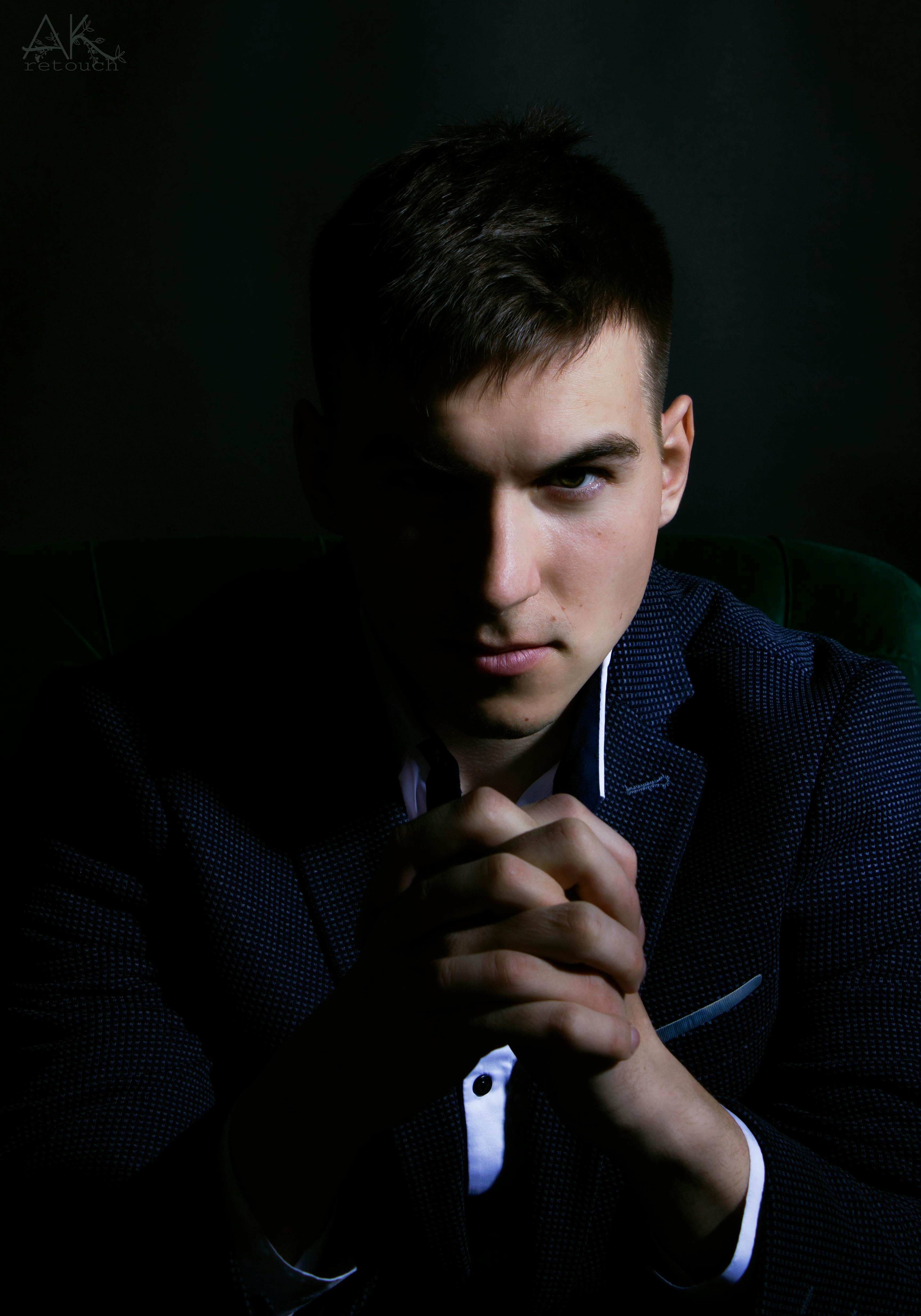 Shadowy portrait of a man in formal attire with a serious expression, hands clasped.