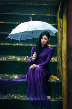 A woman in a purple dress sits on stairs under an umbrella during a rainy day, capturing a serene moment.