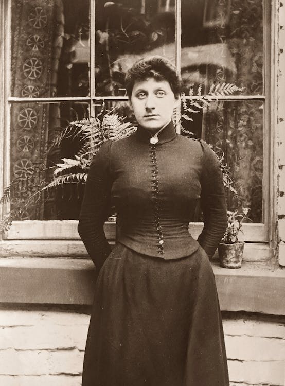 Grayscale Photo of Woman in Long Sleeve Dress Standing Near A Window