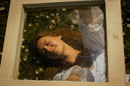 Woman in White Shirt Lying on Green Grass while Holding a Glass Wooden Frame