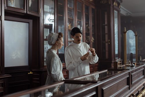 Man and Woman in White Gowns Standing behind the Counter in a Vintage Pharmacy