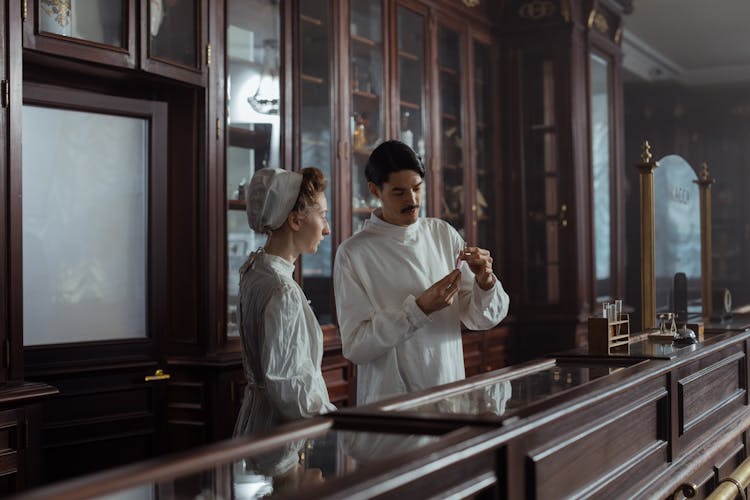 Man And Woman In White Gowns Standing Behind The Counter In A Vintage Pharmacy