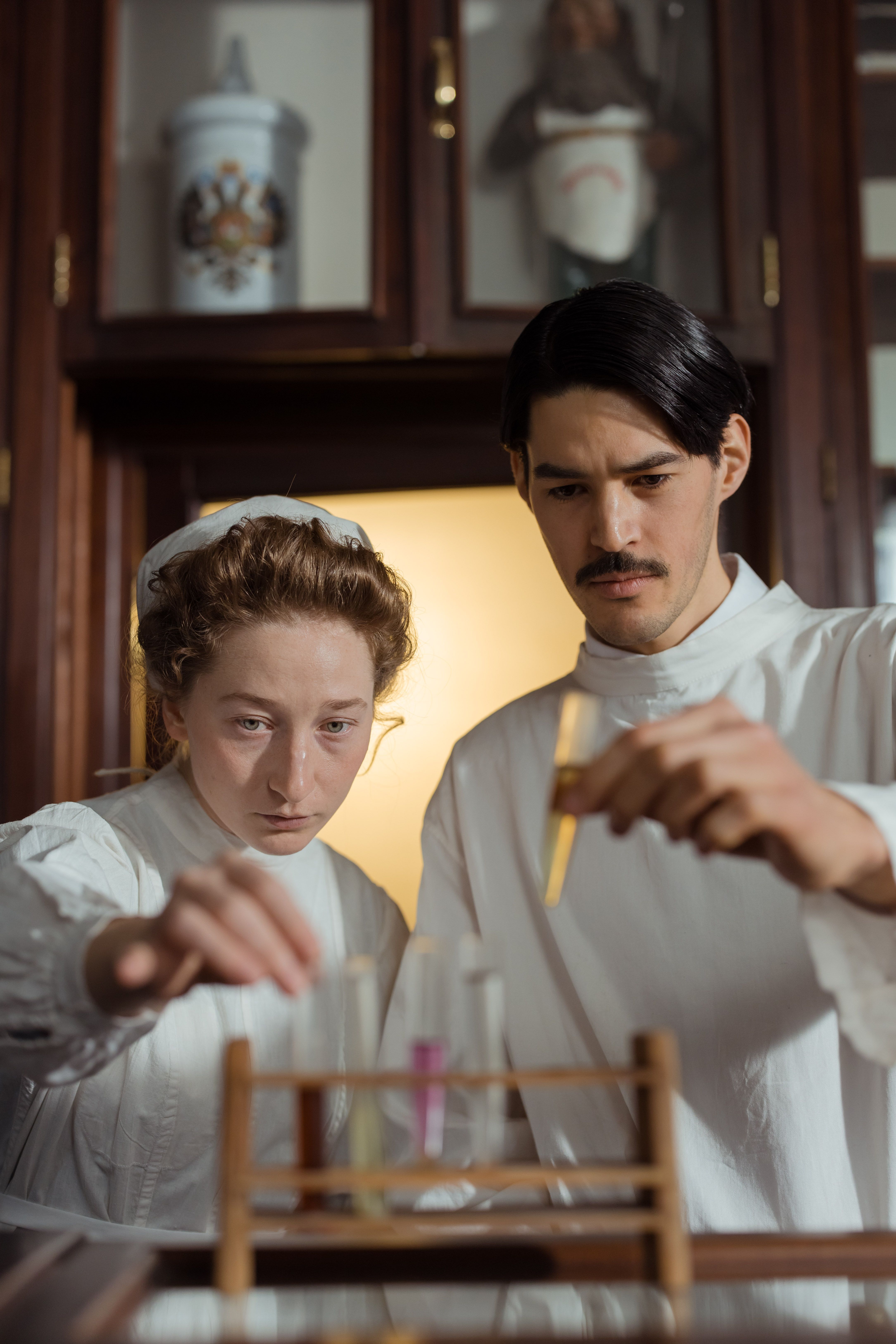 Two pharmacists in historical attire examining test tubes in an old-fashioned laboratory setting.