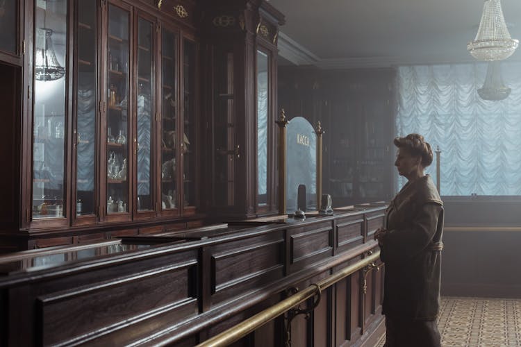 Elderly Woman In Brown Coat Standing By The Counter Of A Pharmacy