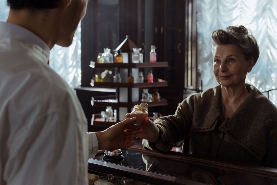 Mulher idosa recebendo um drinque de um atendente em uma loja ou bar com ambiente aconchegante e iluminação natural.