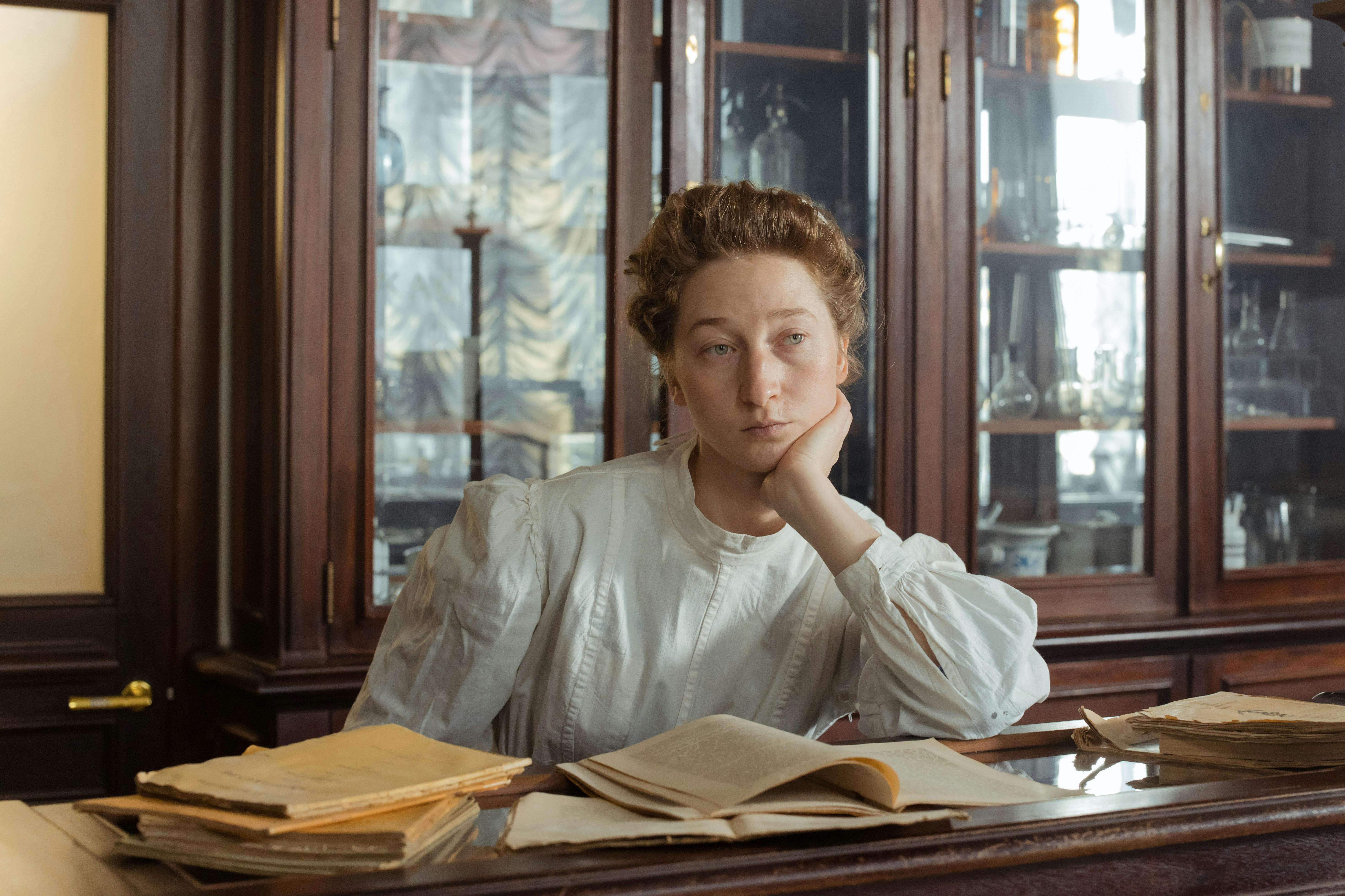 Woman in elegant vintage attire seated in a study room filled with books and scientific instruments.