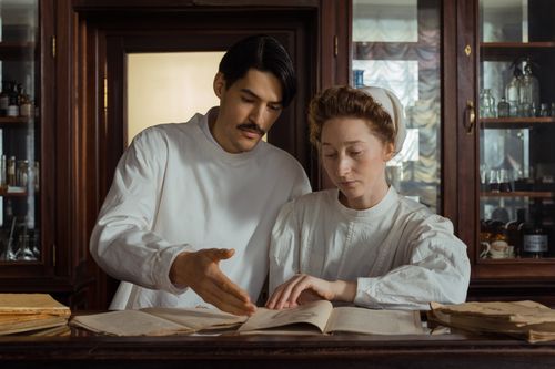 Free Two pharmacists in period attire examine medical texts at a vintage pharmacy counter. Stock Photo