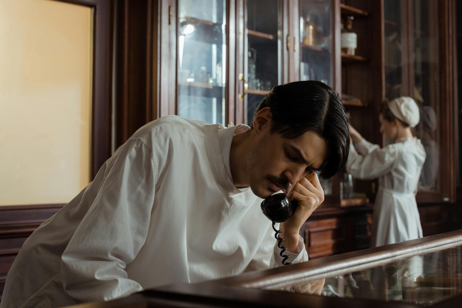 A vintage-style photo showing a pharmacist in conversation on an old-fashioned telephone, with an assistant in the background.