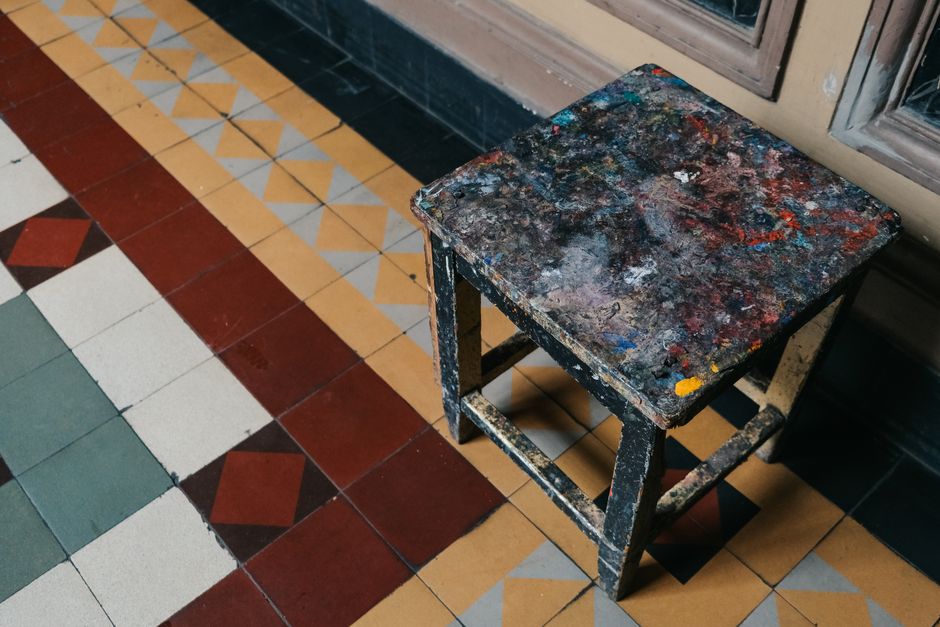 Rustic stool with paint splatters on colorful geometric tiled floor.