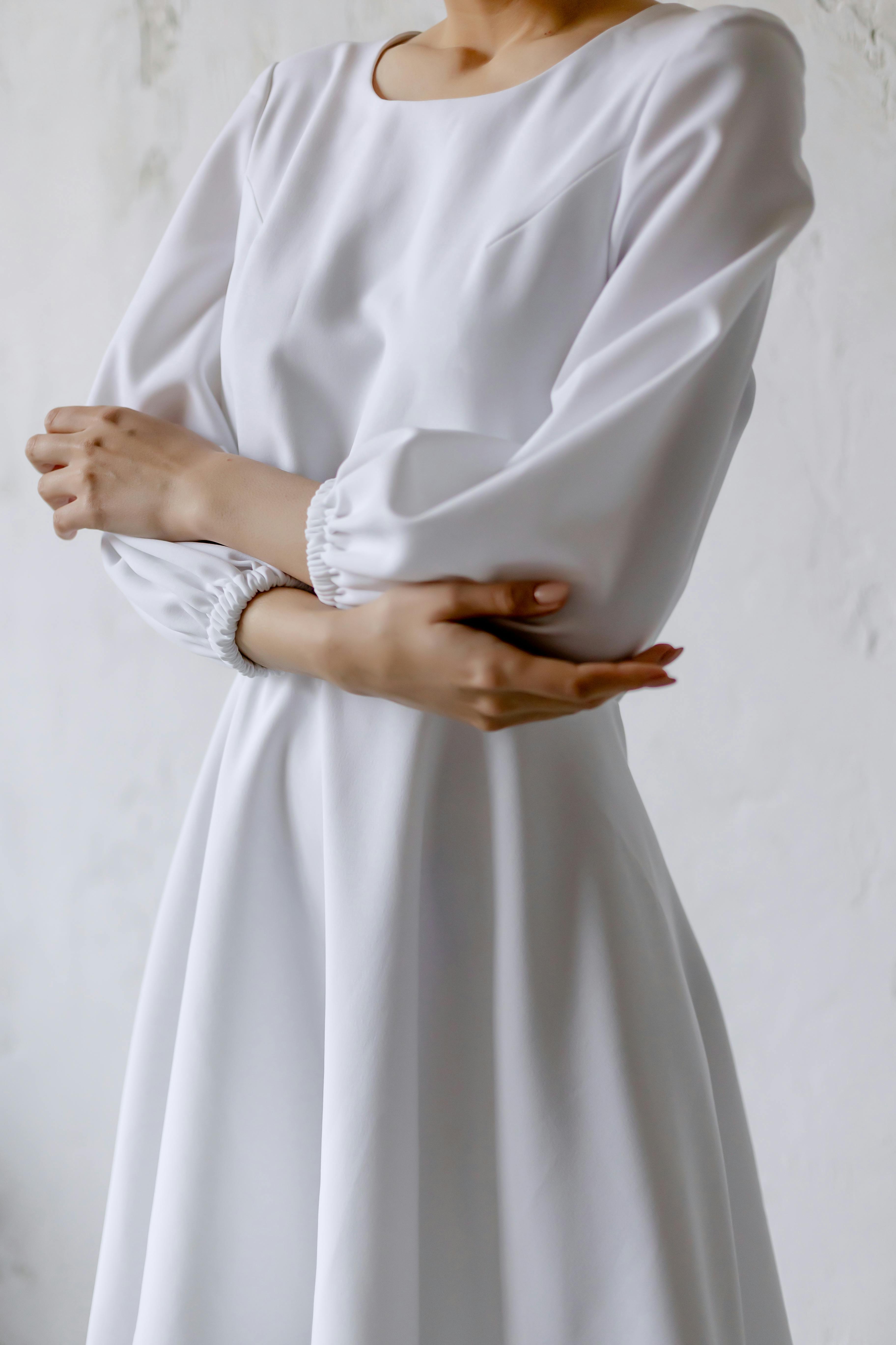 Graceful woman in a minimalist white dress crossing her arms, showcasing elegance and style.