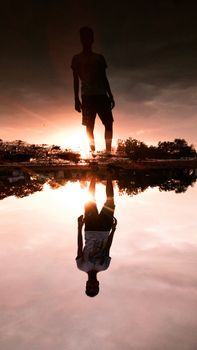 A dramatic silhouette of a man standing by water at sunset with a perfect mirror image reflection.